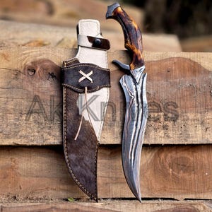 May include: A hunting knife with a patterned steel blade and a brown and tan handle rests beside its leather sheath. The sheath is brown and white with a decorative X stitched on the front. The text "AK Knives" is visible.