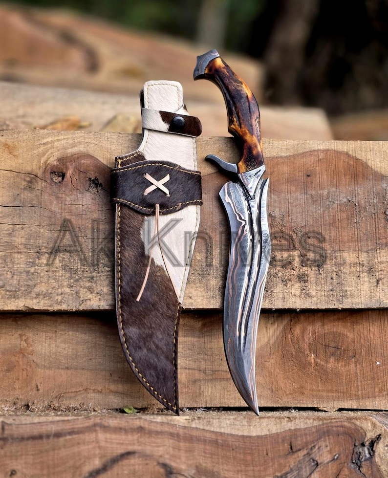 May include: A hunting knife with a patterned steel blade and a brown and tan handle rests beside its leather sheath. The sheath is brown and white with a decorative X stitched on the front. The text "AK Knives" is visible.