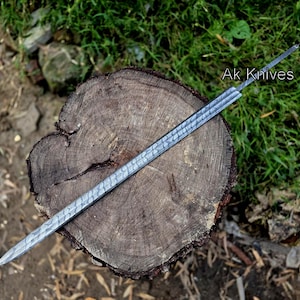 May include: A long, silver-colored knife with a textured blade rests on a wooden tree stump. The knife has a pointed tip and a long, narrow blade. The text "Ak Knives" is visible on the right side of the image.