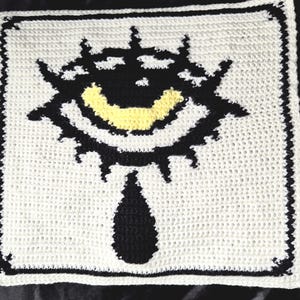 Peut inclure: Pièce carrée en crochet blanc cassé avec un motif d'œil stylisé. L'œil est noir avec un sourire jaune et une larme noire en dessous. La bordure est noire avec un bord festonné.