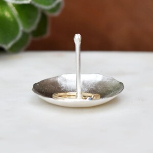 May include: A small, silver-toned ring dish with a central post. The dish has a shallow, leaf-like shape, and holds two rings. The background is blurred, with a succulent plant visible on the left.
