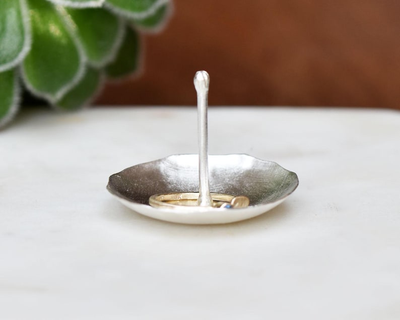 May include: A small, silver-toned ring dish with a central post. The dish has a shallow, leaf-like shape, and holds two rings. The background is blurred, with a succulent plant visible on the left.