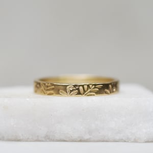 May include: A thin gold band ring with a delicate floral design engraved around the entire band.