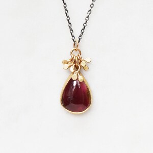 May include: A gold-toned pendant necklace featuring a teardrop-shaped, deep red gemstone. The gemstone is encased in a gold setting and is suspended from a delicate, dark chain. The pendant has a cluster of small gold accents at the top.