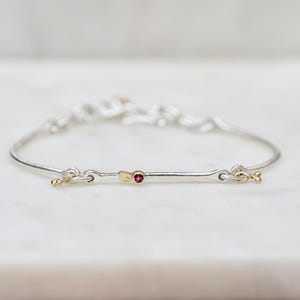 May include: A delicate silver bracelet with gold accents and a small red gemstone. The bracelet features a simple, elegant design with a clasp closure. The jewelry is set against a soft, blurred background.