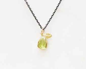 Puede incluir: Collar con colgante dorado en forma de pera, con una piedra preciosa verde claro y una pequeña hoja. El colgante cuelga de una cadena oscura sobre un fondo blanco. La piedra preciosa mide aproximadamente 1,3 cm de largo.