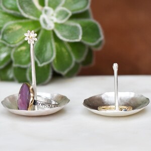 May include: Two silver ring holders with a hammered texture. One has a flower-shaped top, the other a rounded top. Rings with a purple stone and gold band, and a silver band with floral details are displayed.