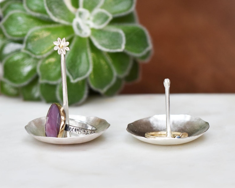 May include: Two silver ring holders with a hammered texture. One has a flower-shaped top, the other a rounded top. Rings with a purple stone and gold band, and a silver band with floral details are displayed.