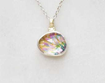 Puede incluir: Un collar de plata con un colgante que presenta un diseño abstracto y colorido. El colgante tiene una fianza dorada y un bisel plateado, con una mezcla de tonos rosa, amarillo, verde y azul. La cadena es delicada y plateada.