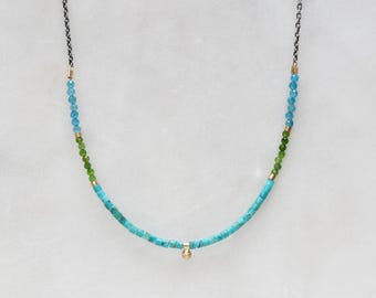 Puede incluir: Un collar de piedras preciosas turquesa, verde y azul con un colgante de oro. El collar está hecho con pequeñas cuentas redondas y está enhebrado en una cadena negra.