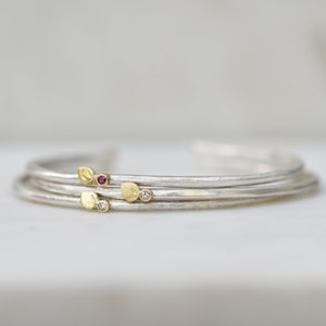 May include: Set of three silver bangles with delicate gold leaf accents and small gemstones. The bangles are stacked on a white surface.