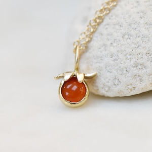May include: A gold-colored necklace featuring a pendant with a round, orange gemstone. The pendant is set in a gold-colored bezel with a floral design. The necklace chain is also gold-colored and delicate.