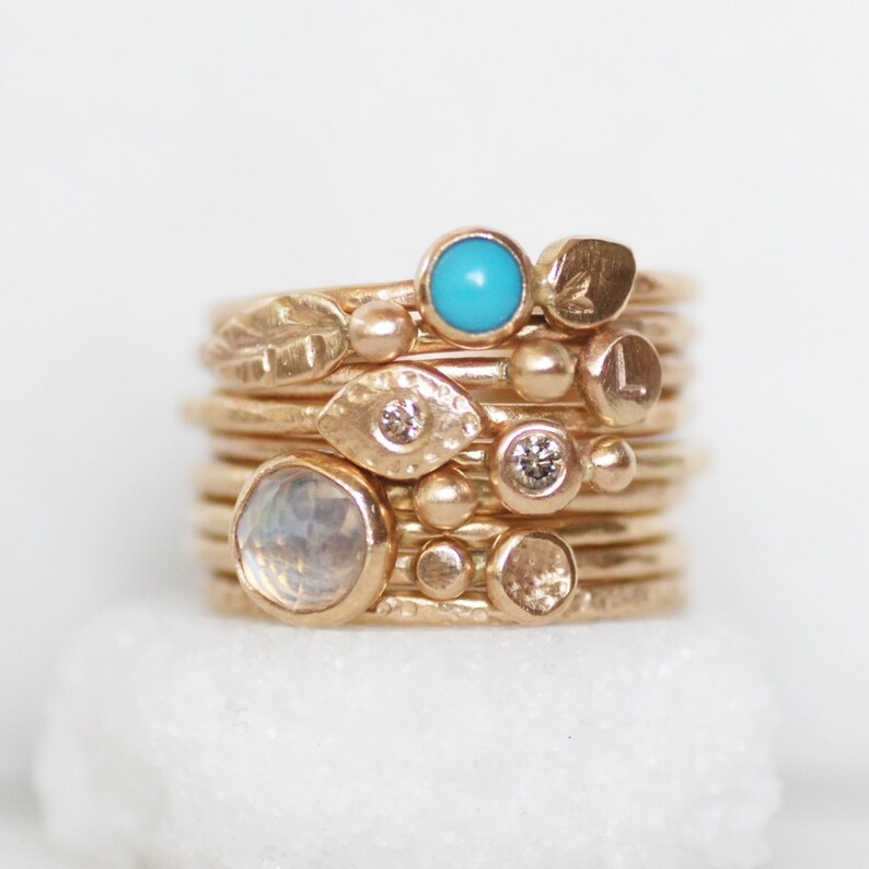 Puede incluir: Una pila de anillos de oro con varias piedras preciosas, incluyendo turquesa, piedra lunar y diamantes. Los anillos son delicados y presentan una variedad de texturas y dise&ntilde;os.