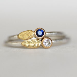 May include: A delicate ring with two small gemstones set in gold. The ring features two gold leaves with a blue sapphire and a white diamond.