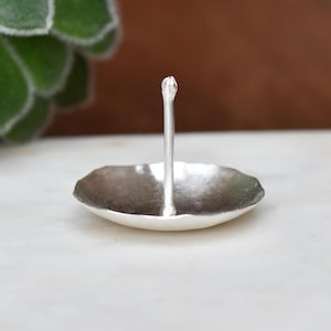 May include: A small, silver-colored ring holder with a textured, shallow dish and a vertical post. The post has a decorative, bud-like top. The ring holder is designed to hold rings and other small jewelry items.