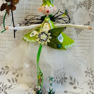 May include: A handcrafted fairy doll featuring a green felt dress, a white tulle skirt, and a floral bodice. The doll has a painted face, pink ears, and a green hat. It holds a brown butterfly wand and has green and white striped legs.