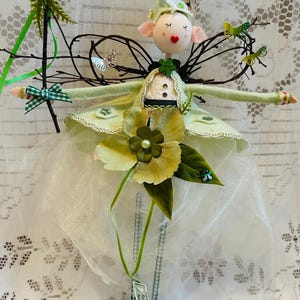 May include: A decorative fairy doll with a painted face, pink ears, and a heart-shaped mouth. The fairy wears a light green dress with a white tulle skirt, adorned with floral accents and butterfly wings. It holds a wand with green ribbons.