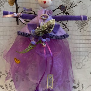 May include: A handcrafted fairy doll in shades of purple, with a felt bodice and a sheer skirt. The doll features a painted face, a floral headdress, and decorative elements like a gold leaf and a small bird. A flower is held in one hand.