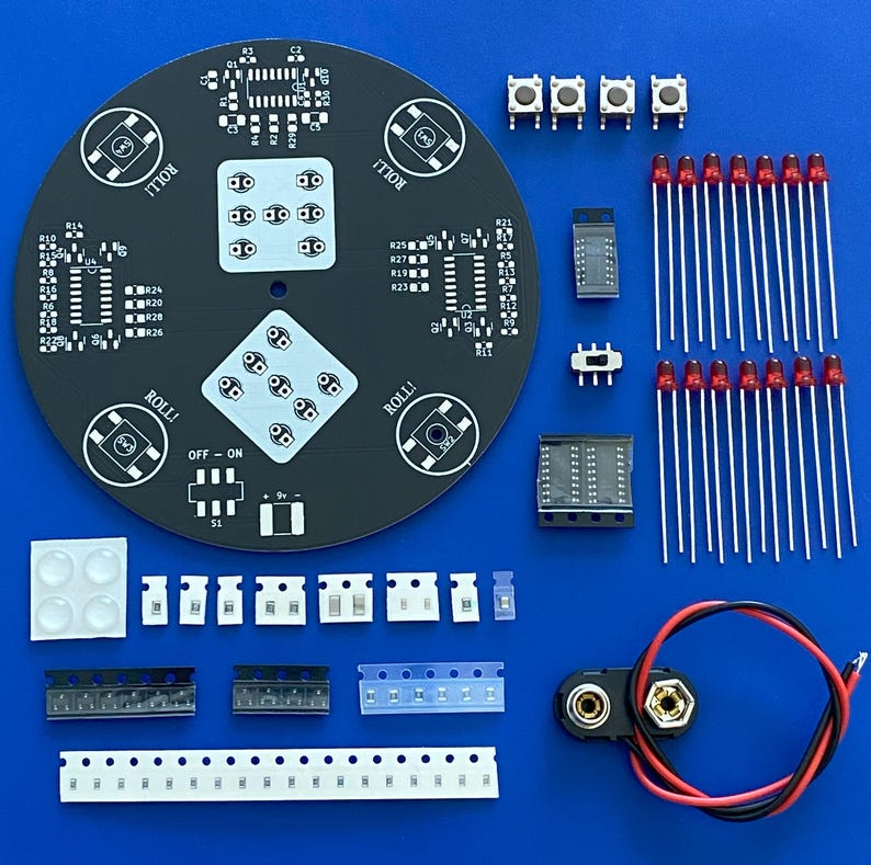 May include: An assortment of electronic components, including a black circuit board with the word "ROLL!" printed on it, LEDs, buttons, and various integrated circuits, arranged on a blue surface. The components are for a DIY electronics project.