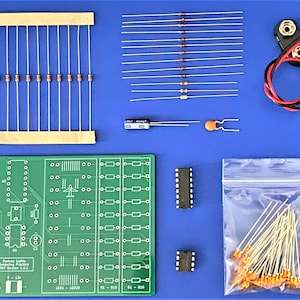 May include: An electronics kit with various components, including a green circuit board, resistors, diodes, and a 9-volt battery connector. The kit also contains integrated circuits and a bag of additional components, all arranged on a blue surface.