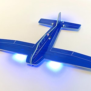 May include: A blue airplane model with white detailing and illuminated blue and red lights. The model has a sleek design with wings and a tail, and is set against a white background.