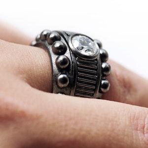 Puede incluir: Un anillo de plata con una gran piedra preciosa transparente engastada en una banda texturizada. La banda está decorada con pequeñas cuentas de plata.