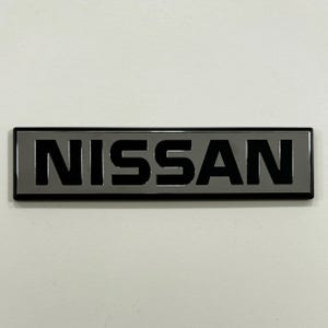 Puede incluir: Un emblema rectangular de Nissan en negro y gris. La palabra "NISSAN" se muestra en letras negras y en negrita sobre un fondo gris. El emblema tiene un acabado brillante y está sobre un fondo blanco.