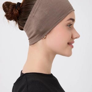 May include: A taupe-colored headband worn by a person with brown hair styled in a bun. The headband is made of a soft, stretchy material and is designed to keep hair out of the face. The person is wearing a black top.