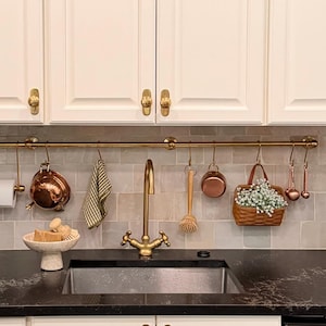 Może przedstawiać: Jasna kuchnia z białymi szafkami i złotymi uchwytami. Zlew i kran ze stali nierdzewnej są wyśrodkowane pod mosiężnym prętem z przyborami kuchennymi. Wazon z białymi kwiatami stoi na czarnym blacie.