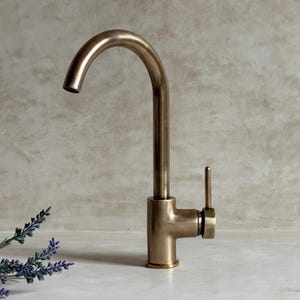 May include: A bronze-colored kitchen faucet with a curved spout and a single lever handle. The faucet has a tall, slender design and is mounted on a white surface. The background is a neutral, textured wall.