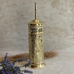 Puede incluir: Un recipiente alto y ornamentado de metal dorado con tapa y asa. El recipiente presenta intrincados patrones en relieve y una banda calada decorativa cerca de la parte superior. El dise&ntilde;o sugiere un prop&oacute;sito decorativo o ceremonial.