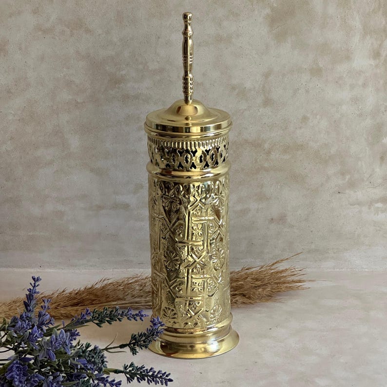 Puede incluir: Un recipiente alto y ornamentado de metal dorado con tapa y asa. El recipiente presenta intrincados patrones en relieve y una banda calada decorativa cerca de la parte superior. El dise&ntilde;o sugiere un prop&oacute;sito decorativo o ceremonial.