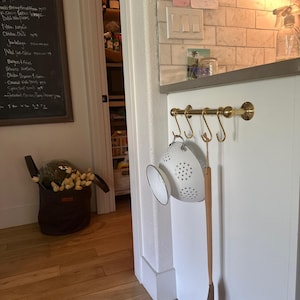 May include: A kitchen scene with a brass-toned hanging rack holding a white colander and a wooden spatula. A dark brown tote bag filled with yellow flowers sits on the light-colored hardwood floor. A chalkboard menu is visible.