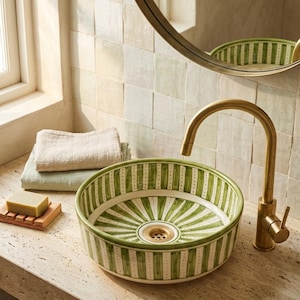 Peut inclure: Une vasque ronde en céramique ornée d'un motif rayé peint à la main en vert et blanc avec une bonde centrale. La vasque est accompagnée d'un robinet en laiton haut et courbé. Deux serviettes en lin pliées et un savon sur un plateau en bois.