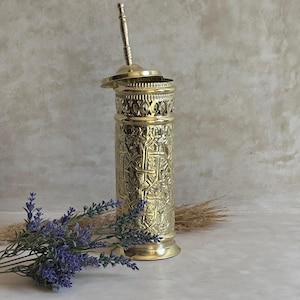 Puede incluir: Un recipiente de metal dorado y ornamentado con tapa y asa. El recipiente presenta intrincados patrones en relieve y una secci&oacute;n calada decorativa cerca de la parte superior. Una ramita de lavanda y hierba seca est&aacute;n en primer plano.