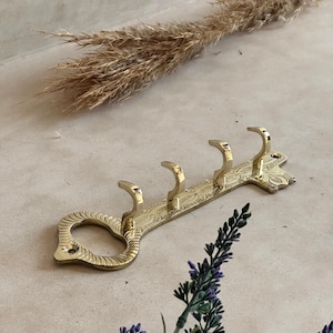 May include: A gold-colored metal key-shaped wall hook with four curved hooks. The hook has an ornate, engraved design and a decorative keyhole shape at the end. The hook is designed to hang items.