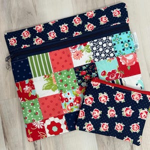 May include: Two quilted fabric pouches. The larger pouch has a patchwork design with red, green, white, and blue floral patterns. The smaller pouch is navy blue with a red zipper and a repeating pattern of pink roses. Both have zippers.