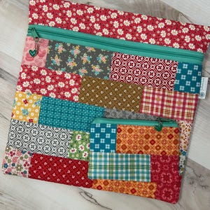 May include: A large rectangular zippered pouch and a smaller matching zippered pouch, both featuring a patchwork design of various floral, geometric, and plaid fabric patterns in red, teal, yellow, and gray, with teal zippers and heart charms.