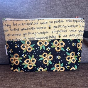 May include: A rectangular fabric pouch with a black handle and a zipper closure. The top section is a light yellow color with inspirational phrases and bee graphics. The bottom section features a black background with sunflowers and bees.
