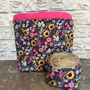 May include: Two fabric storage baskets. The larger basket has a dark blue floral pattern with yellow sunflowers, pink and white flowers, and a bright pink interior. The smaller basket has the same floral pattern and holds a ball of twine.