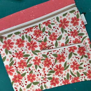 May include: Two zippered pouches with a floral pattern. The pouches feature a white background with red flowers, green leaves, and small red hearts. The top of the pouches are a solid coral color with a white polka dot pattern. The zippers are metallic.