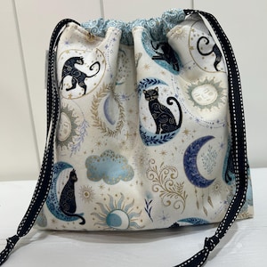 May include: A drawstring pouch made of cream-colored fabric featuring a repeating pattern of black cats sitting on blue crescent moons, golden suns, and light blue clouds. The bag is cinched with black ribbon drawstrings with white dots.