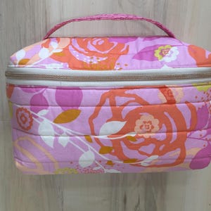 May include: A quilted cosmetic bag with a floral pattern in shades of pink, orange, and white. The bag has a rectangular shape, a zipper closure, and a small handle. The bag is approximately 8 inches long, 6 inches wide, and 5 inches tall.