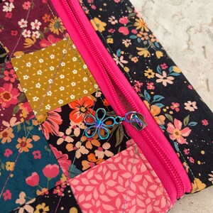 May include: Close-up of a quilted floral fabric pouch with a bright pink zipper. The fabric features a patchwork design with various floral patterns in shades of pink, orange, yellow, and black. The zipper pull is a flower-shaped charm.