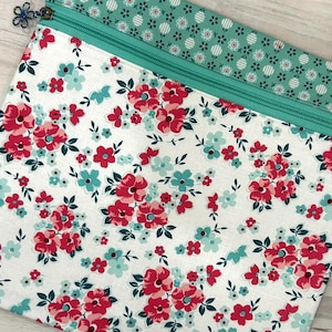 May include: A fabric pouch featuring a white background with a floral pattern of pink and light blue flowers and dark green leaves. The top section has a teal zipper and a teal fabric border with a small white circular pattern and a flower charm.