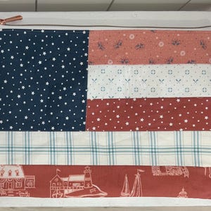 May include: A rectangular fabric zipper pouch featuring a patchwork design. The left side is navy blue with white stars. The right side consists of horizontal stripes in coral, white with nautical icons, red with white stars, white with blue plaid, and coral with lighthouse illustrations.
