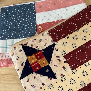 May include: Two rectangular fabric pouches featuring patchwork designs. The top pouch has a blue section with white stars and pink striped sections. The bottom pouch features a large dark blue star, red and yellow squares, and cream stripes.