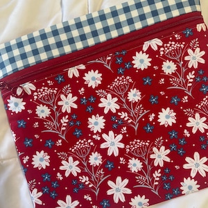 May include: A rectangular zippered pouch featuring a red fabric body with a white and blue floral pattern. The top section of the pouch is accented with a blue and white gingham check fabric. A red zipper runs horizontally across the top.