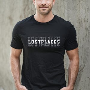 Könnte beinhalten: Schwarzes T-Shirt mit dem Text "LOSTPLACES" in Weiß, dreifach gestapelt. Das Shirt hat kurze Ärmel und scheint aus einem weichen Material zu sein. Die Person, die das Shirt trägt, steht vor einer grauen Wand.