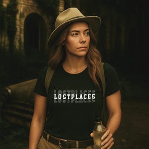 Könnte beinhalten: Eine Person trägt einen khakifarbenen Fedora-Hut und ein schwarzes T-Shirt mit dem weißen Text "LOSTPLACES". Sie trägt auch einen Rucksack und hält eine klare Flasche. Der Hintergrund zeigt eine alte Steinstruktur und üppiges Grün.